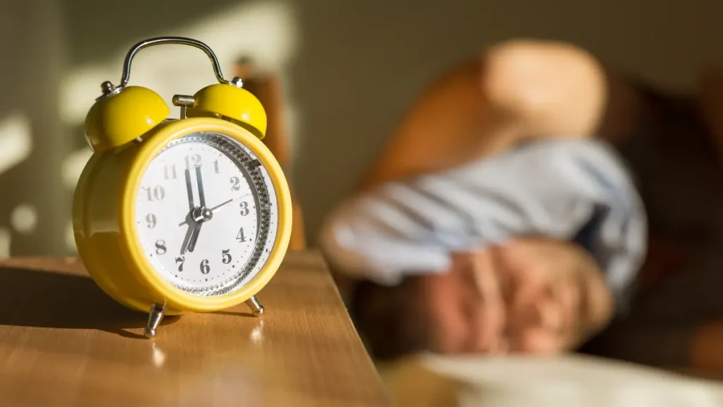 A yellow alarm clock on a wooden table with a blurred man waking up in the background.