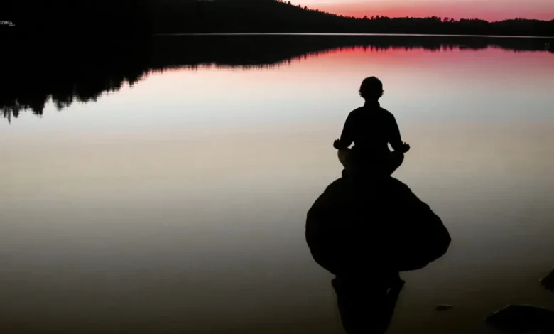 Silhouette of a person meditating on a rock in a calm lake during sunset.