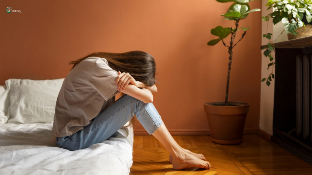 Sad woman sitting on the floor with head down, leaning against a bed in a cozy room