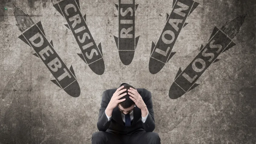 A stressed businessman sits with his head in his hands as missiles labeled "Debt," "Crisis," "Risk," "Loan," and "Loss" approach.